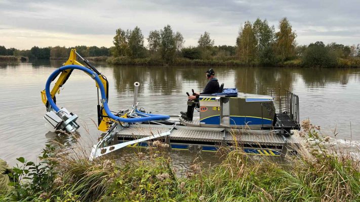 Seitenansicht einer Gewässerreinigungsmaschine welche ein paar Meter vom Ufer durch einen Fluss fährt und Verunreinigungen entfernt.